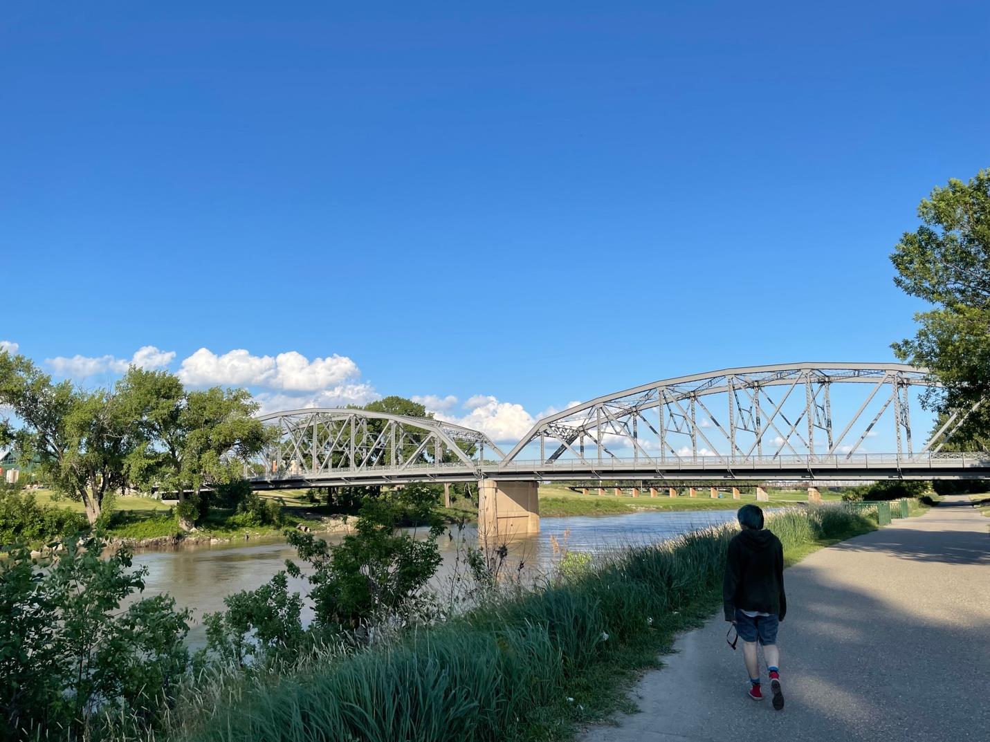 One of the bridges over the Red River in Grand Forks, ND. Behind the bridge is a brilliant blue sky with a few clouds hovering nearer the horizon. In the foreground, a person is walking along a path toward the bridge.