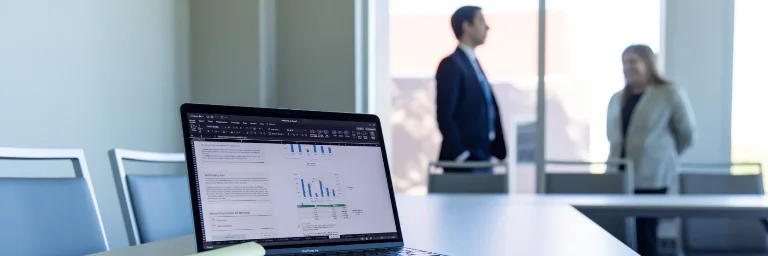 A laptop screen shows financial data related to accounting, while two professionals engage in conversation in the background.