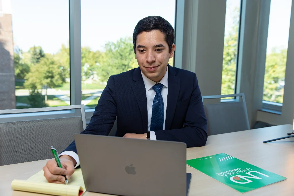 A focused male accountant works on his laptop, simultaneously transcribing notes in his notepad.