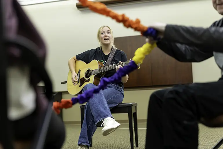 Aubrey plays a guitar as two people move in front